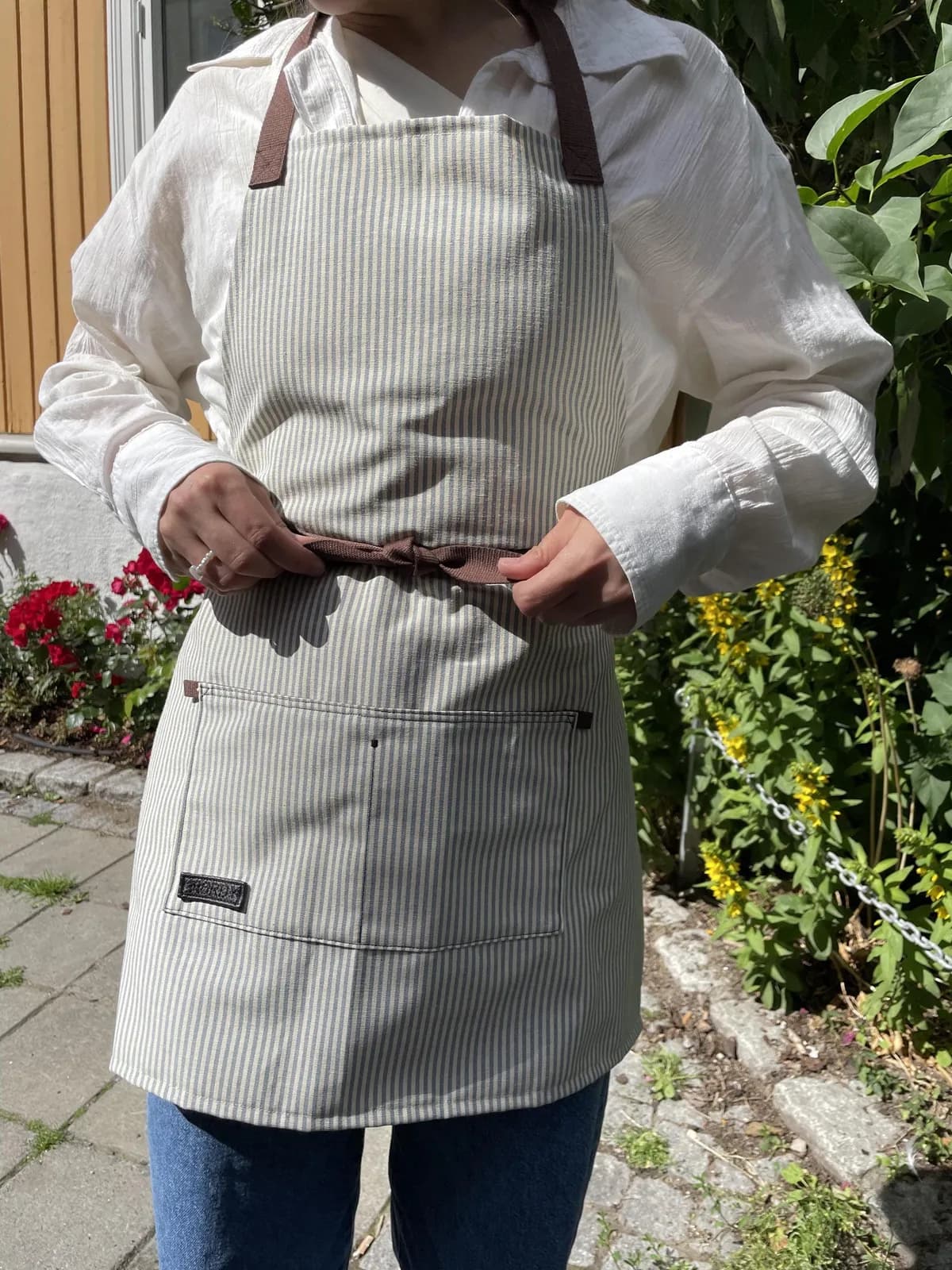 Grått forkle med brodert logo, båret i en hage